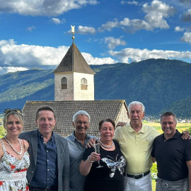 Sechs Personen vor einer Kirche in den Bergen, mit einem Glas Sekt in der Hand.
